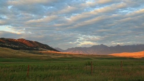 A scenic landscape featuring rolling green fields, distant mountains, and a dramatic sky with scattered clouds at sunset.