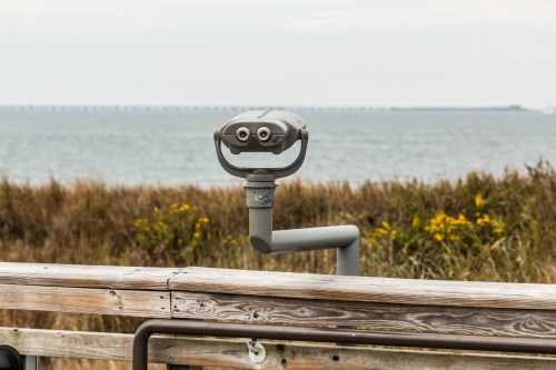 A coin-operated binocular viewer on a wooden railing overlooking a calm body of water and distant shoreline.