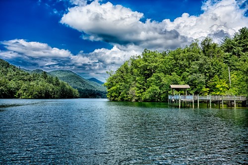 A serene lake surrounded by lush green trees and mountains, with a wooden dock extending into the water under a blue sky.