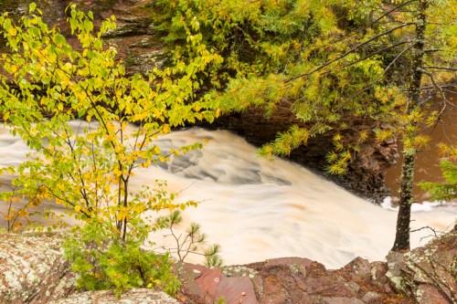 A flowing river surrounded by lush green trees and autumn leaves, cascading over rocky terrain.