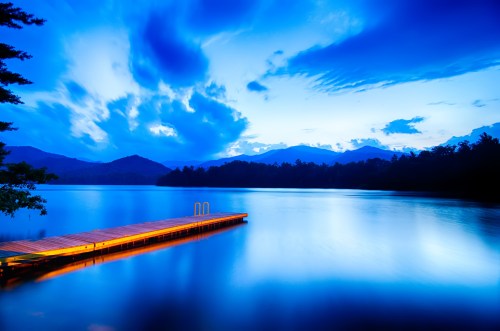 A serene lake at dusk with a wooden dock extending into calm waters, surrounded by mountains and dramatic clouds.