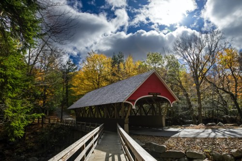 A red covered bridge surrounded by autumn trees under a cloudy sky.