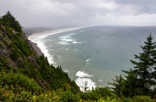 A coastal view featuring a rocky cliff, lush greenery, and waves crashing on a sandy beach under a cloudy sky.
