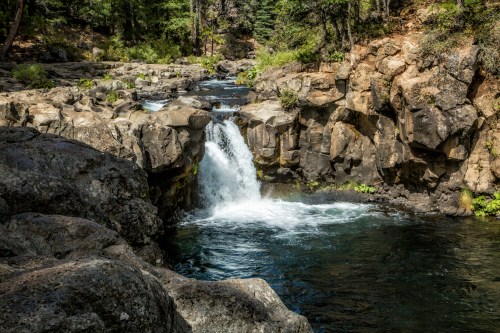 A serene waterfall cascades over rocky terrain, surrounded by lush greenery and clear blue water.