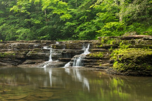 A serene waterfall cascades over rocky ledges into a calm pool, surrounded by lush green trees and foliage.