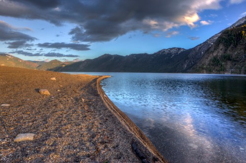 A serene lakeside view with a sandy shore, mountains in the background, and a cloudy sky reflecting on the water.