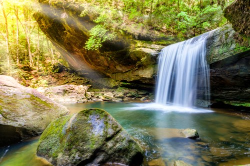 A serene waterfall cascades over rocks into a tranquil pool, surrounded by lush green trees and sunlight filtering through.
