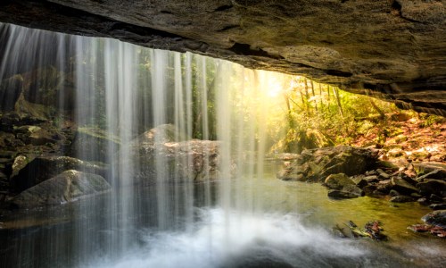 A serene waterfall cascades over rocks, surrounded by lush greenery and soft sunlight filtering through the trees.