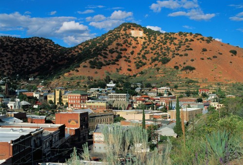 A scenic view of a historic town nestled in a valley, surrounded by mountains and blue skies with scattered clouds.