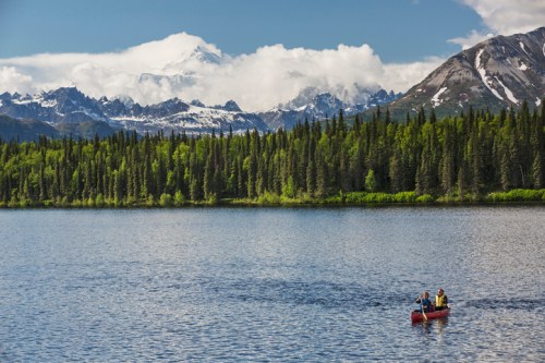A canoeist paddles on a serene lake surrounded by lush forests and snow-capped mountains under a blue sky.
