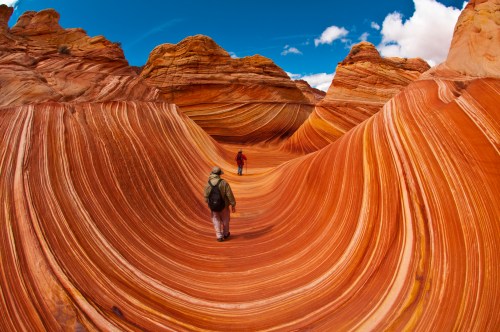 Two hikers explore the colorful, wavy rock formations of The Wave in Arizona under a bright blue sky.