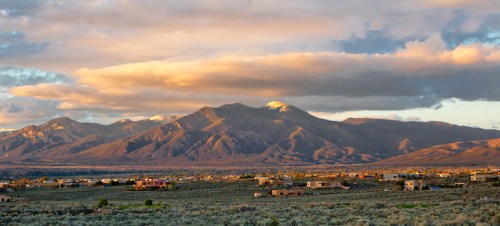 A panoramic view of mountains under a colorful sky, with a valley and scattered buildings in the foreground.