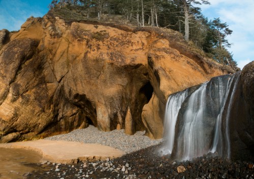 A serene waterfall cascades over rocky cliffs, surrounded by lush greenery and a sandy beach.