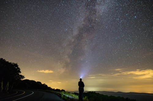 A person stands on a road at night, illuminating the sky with a flashlight, surrounded by a starry Milky Way backdrop.
