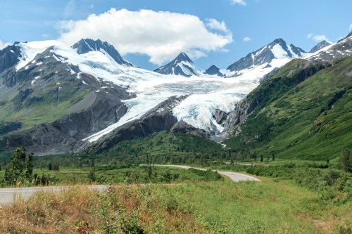 A scenic view of snow-capped mountains and a glacier, with a winding road and lush greenery in the foreground.