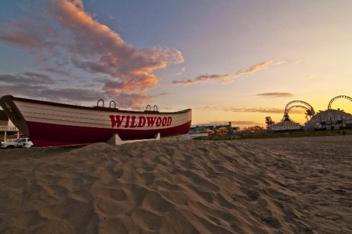 A large boat labeled "WILDWOOD" on sandy beach with a sunset and amusement park rides in the background.