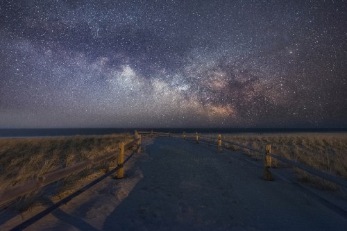 A sandy path leads to the ocean under a starry night sky filled with the Milky Way. Fences line the path.