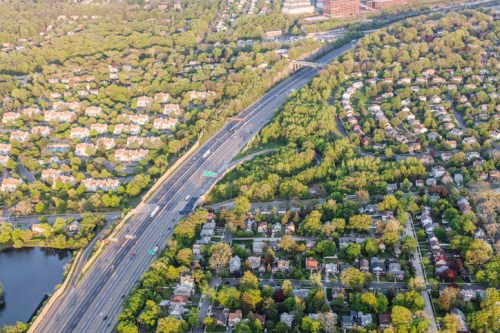 Aerial view of a highway surrounded by lush greenery and residential areas, with a river visible nearby.