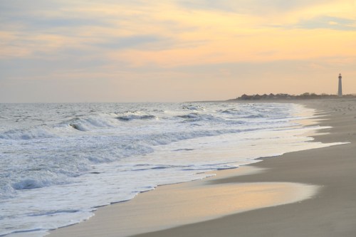 A serene beach at sunset, with gentle waves and a distant lighthouse against a pastel sky.