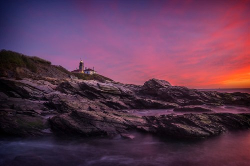 A lighthouse on rocky shore at sunset, with vibrant pink and purple skies reflecting over calm waters.