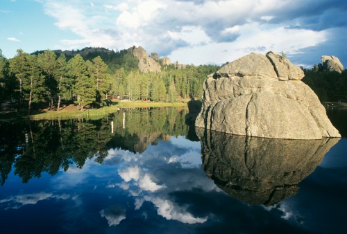 A serene lake reflects a large rock and trees under a cloudy sky, with distant rocky formations in the background.