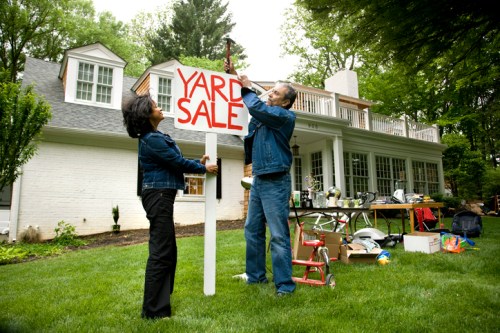A man and woman set up a yard sale sign in front of a house, surrounded by various items for sale on the lawn.