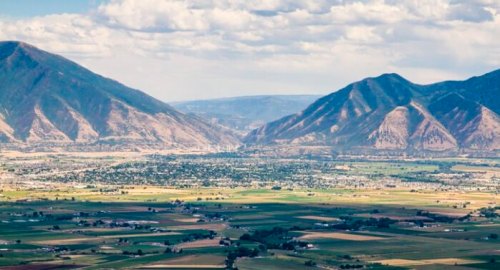 A panoramic view of a valley surrounded by mountains, with fields and a small town visible below under a cloudy sky.