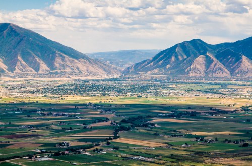 A panoramic view of green fields and valleys surrounded by mountains under a partly cloudy sky.