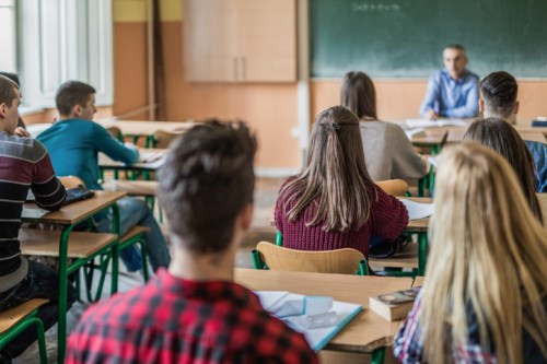 A classroom scene with students seated at desks, facing a teacher at the front near a chalkboard.