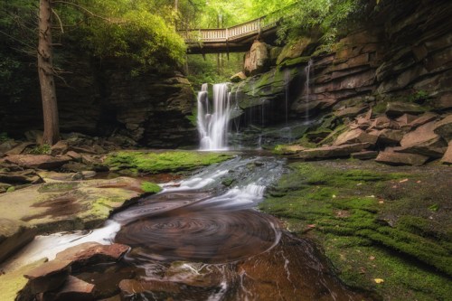 A serene waterfall cascades over rocks, surrounded by lush greenery and a wooden bridge above.