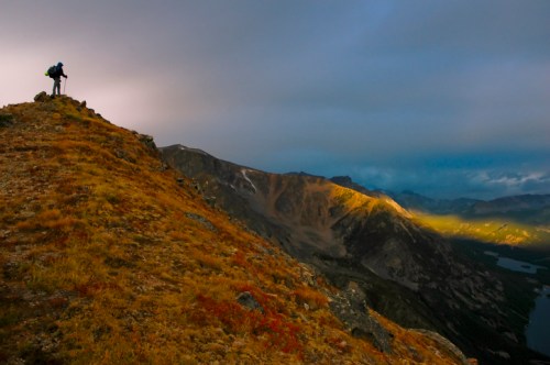 A hiker stands on a rocky ridge, overlooking a valley with mountains and a lake, illuminated by soft sunlight.