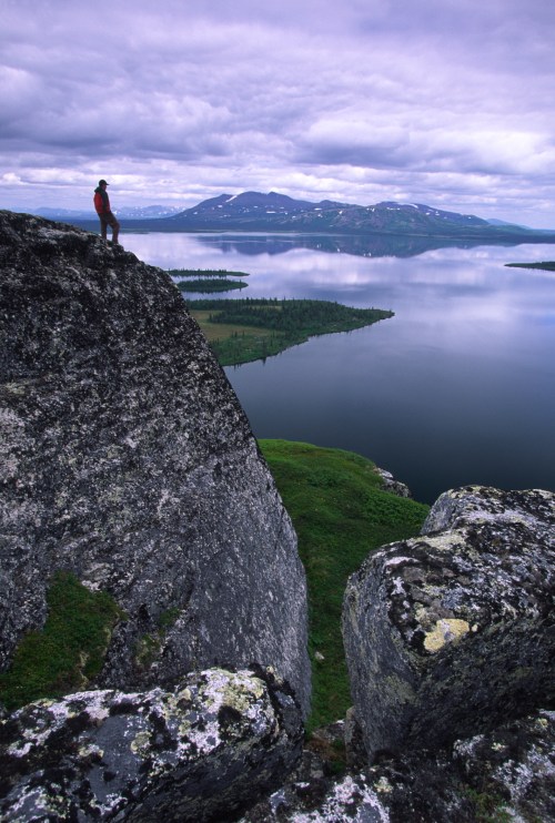 A person stands on a rocky ledge overlooking a calm lake and distant mountains under a cloudy sky.