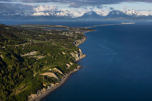 Aerial view of a coastal landscape with mountains in the background and a calm sea, featuring lush greenery along the shore.