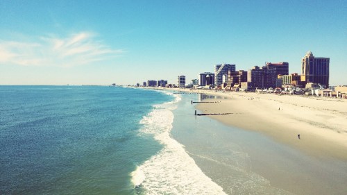 A scenic view of a beach with gentle waves, stretching towards a skyline of buildings under a clear blue sky.