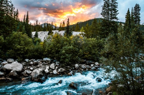 A serene river flows through lush greenery, with a vibrant sunset illuminating the sky and mountains in the background.
