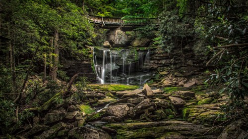 A serene waterfall cascades over rocks, surrounded by lush greenery and a wooden bridge above.