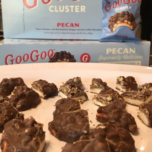A plate of GooGoo Clusters with pecans, surrounded by boxes of the candy in the background.