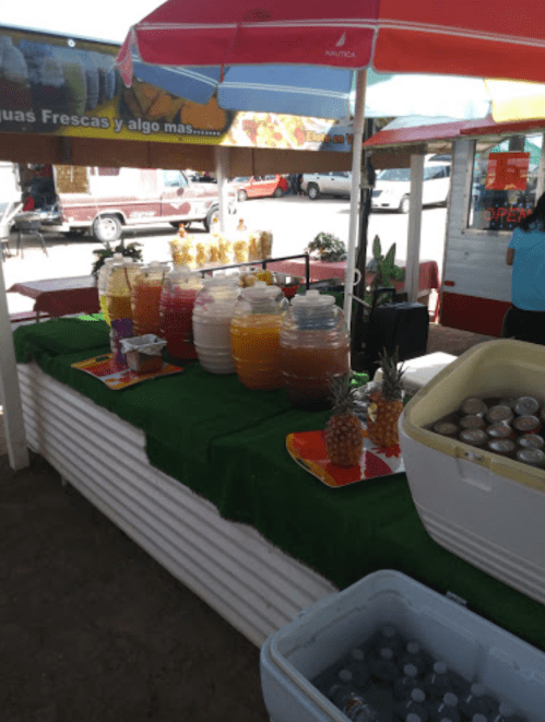 A colorful stand displaying various drinks in jars, with pineapples and a cooler in a sunny outdoor setting.