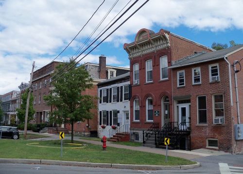 A street view of historic brick and wooden houses with a tree and a fire hydrant in the foreground under a cloudy sky.