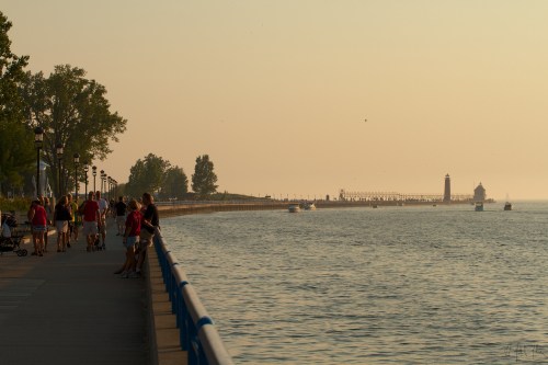 A serene waterfront scene at sunset, with people walking along a path and a lighthouse in the distance.