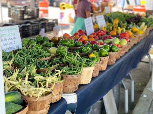 A vibrant farmers market display featuring baskets of fresh green beans, peppers, cucumbers, and various vegetables.