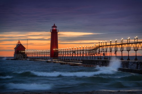 A red lighthouse stands on a pier at sunset, with waves crashing against the shore and a colorful sky in the background.