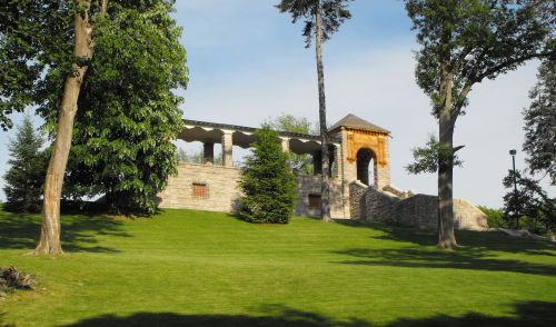 A stone structure with columns and a decorative arch, surrounded by lush green grass and trees under a clear blue sky.