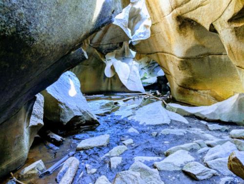 A rocky cave with smooth stone walls and a small stream running through, illuminated by natural light.