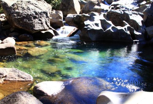 A serene pool of clear water surrounded by large rocks and gentle flowing water, reflecting sunlight.