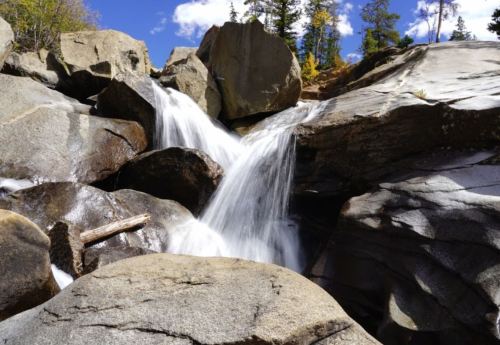A serene waterfall cascading over large rocks, surrounded by trees and a clear blue sky.