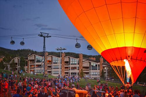 A vibrant hot air balloon inflates at dusk, surrounded by a crowd and scenic buildings, with gondolas overhead.