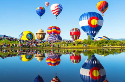 Colorful hot air balloons float above a crowd by a reflective lake under a clear blue sky.