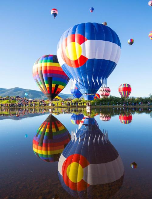 Colorful hot air balloons reflect in a calm lake, with a clear blue sky and mountains in the background.