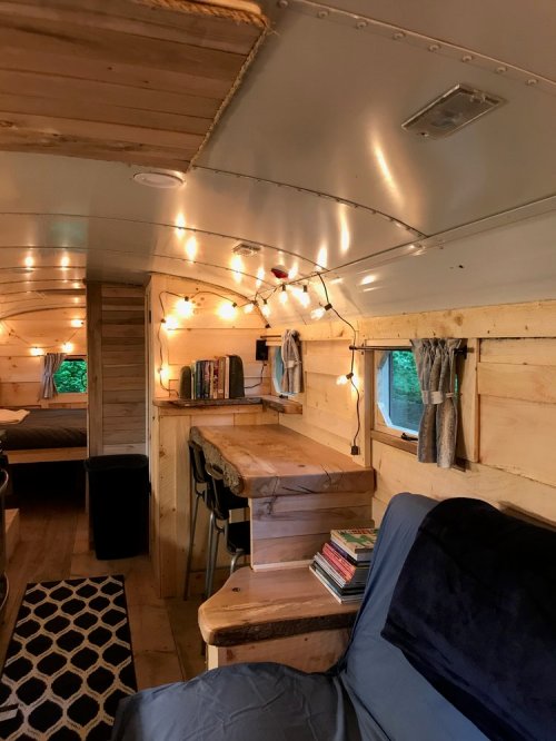 Cozy interior of a converted bus with wooden walls, string lights, a dining table, and a seating area.
